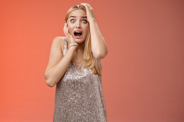 Shocked terrified blond woman horrified see crime screaming pop eyes shouting hold hands head afraid trembling fear standing frightened drop jaw gasping face terrible accident, red background.