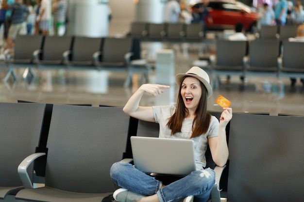 Shocked traveler tourist woman in hat sit with crossed legs with laptop pointing finger on credit card wait in lobby hall at airport