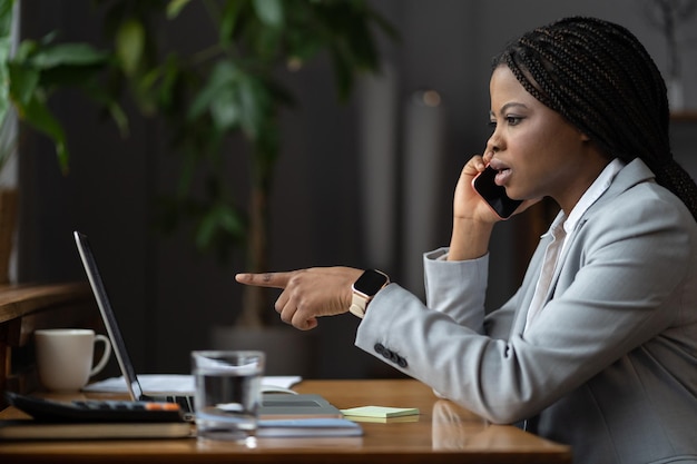 Shocked unhappy afro businesswoman talk on phone call point finger at laptop screen solving problem