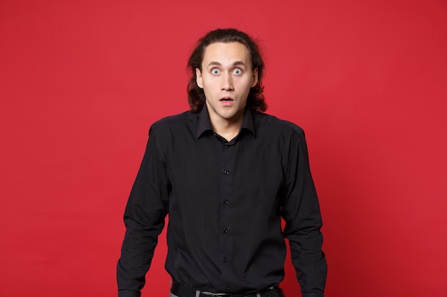 Shocked upset stylish young curly long haired man in black shirt posing isolated on red wall background studio portrait. People sincere emotions lifestyle concept. Mock up copy space. Looking camera.