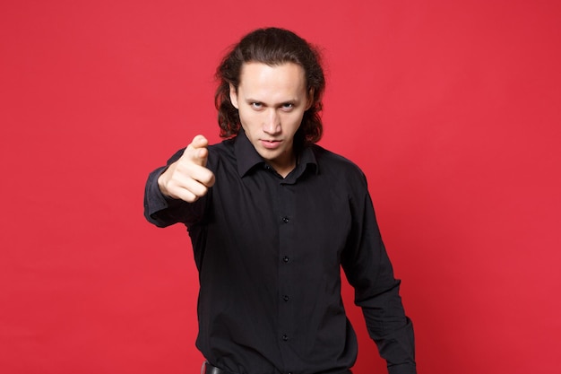 Shocked upset stylish young curly long haired man in black shirt posing isolated on red wall background studio portrait. People sincere emotions lifestyle concept. Mock up copy space. Looking camera.