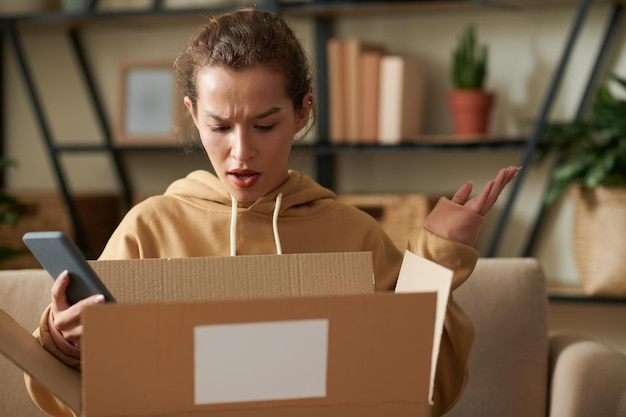 Photo shocked woman holding mobile phone and opening the cardboard box she displeased with delivery of her parcel