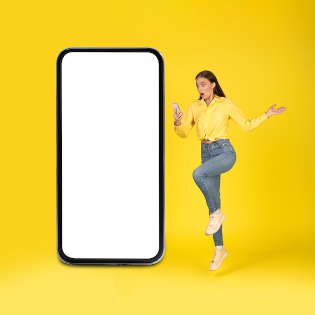 Shocked woman holding phone posing near huge smartphone yellow background