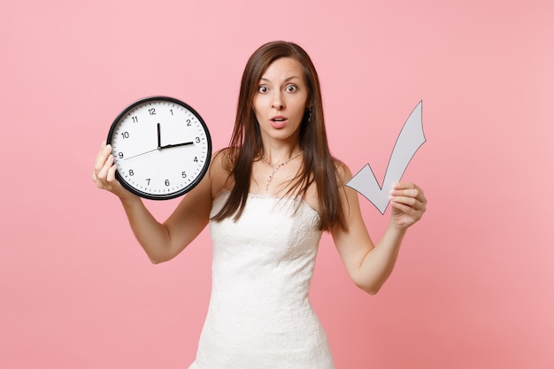 Shocked woman in lace white dress holding check mark and round alarm clock