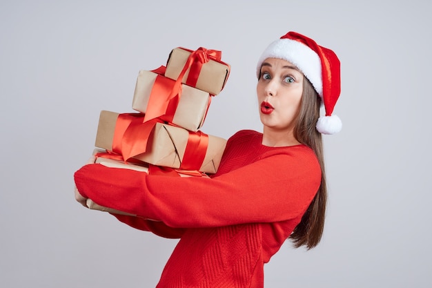 Shocked woman in santa hat, red sweater, holding a bunch of gifts with a red ribbon. Holiday concept, Christmas, New Year.