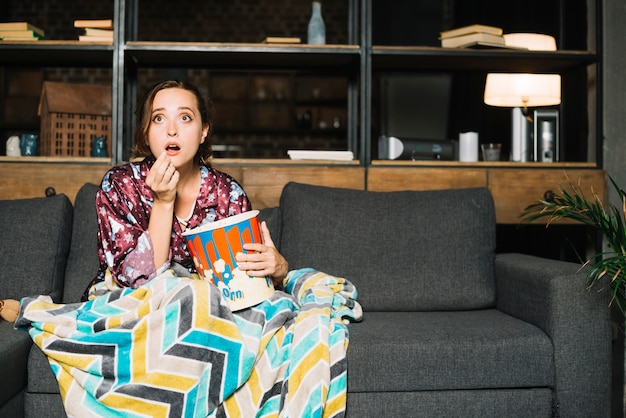 Shocked woman sitting on sofa with popcorn watching television