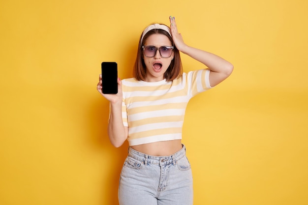 Shocked woman wearing striped t shirt and jeans posing isolated over yellow background standing smart phone showing empty display with copy space for promotional test expressing astonishment