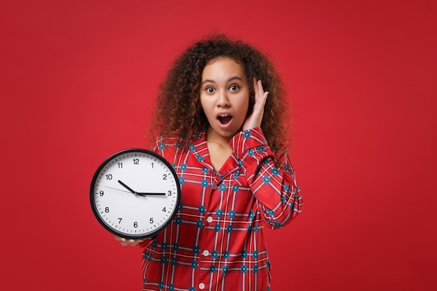 Shocked young african american girl in pajamas homewear posing while resting at home isolated on red background. Relax good mood lifestyle concept. Mock up copy space. Hold clock put hand on cheek.