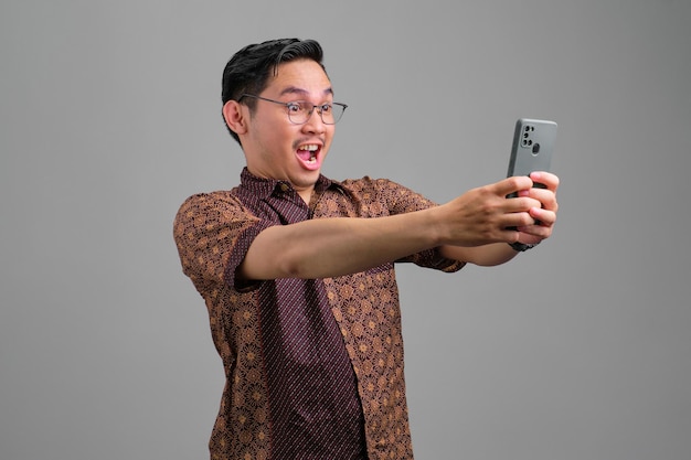 Shocked young Asian man wearing batik shirt looking at smartphone with stunned facial expression isolated on grey background