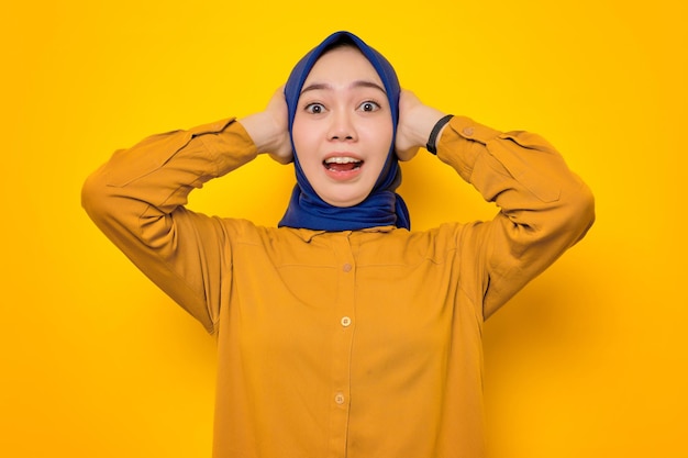 Shocked young Asian Muslim woman dressed in orange shirt looking at camera with open mouth and grabbing head isolated on yellow background