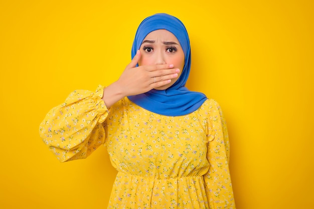 Shocked young Asian woman in casual dress covering mouth with hand isolated on yellow background