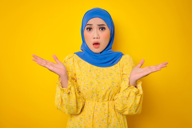 Shocked young Asian woman in casual dress looking at camera with opened mouth spreading hands isolated on yellow background