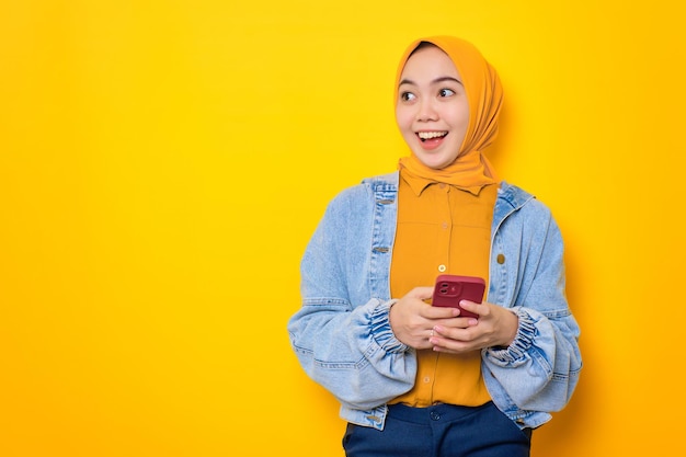 Shocked young Asian woman in jeans jacket holding mobile phone and looking aside at copy space isolated over yellow background