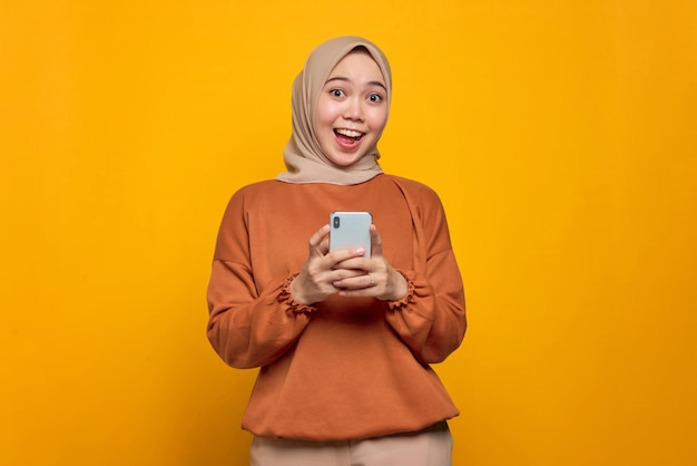 Shocked young Asian woman in orange shirt using mobile phone with open mouth received good news isolated over yellow background