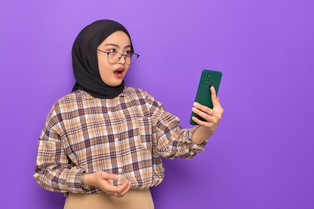 Shocked young Asian woman in plaid shirt holding a mobile phone looking aside at copy space isolated on purple background