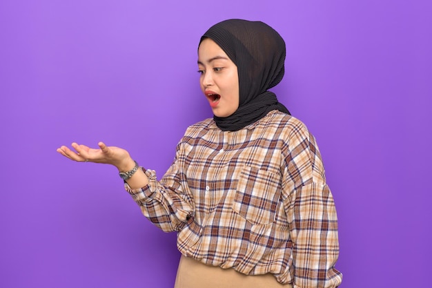 Shocked young Asian woman in plaid shirt showing copy space on her palm isolated on purple background