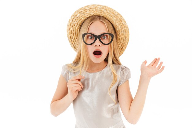 Shocked young blonde girl in dress and straw hat holding fake eyeglasses on white