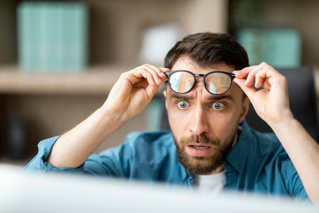 Shocked young businessman taking off glasses and looking at laptop screen