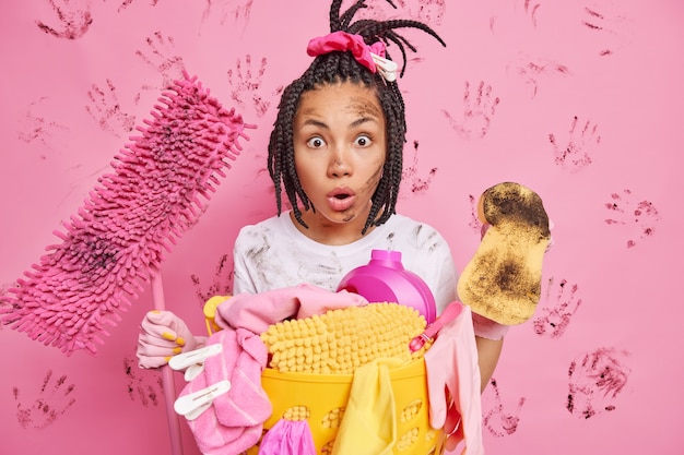 Shocked young ethnic housemaid looks at very dirty room with chaos around holds sponge and mop busy tidying up kitchen collects laundry in basket does house chores poses against pink wall