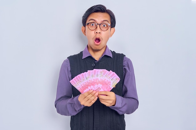 Shocked young handsome Asian man in shirt clothes holding cash money in rupiah banknotes and looking at camera isolated on white background People lifestyle concept