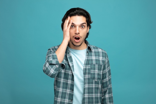 shocked young handsome man keeping hand on head looking at camera isolated on blue background