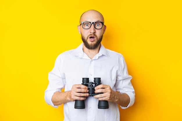 Shocked young man in glasses holding binoculars on yellow background