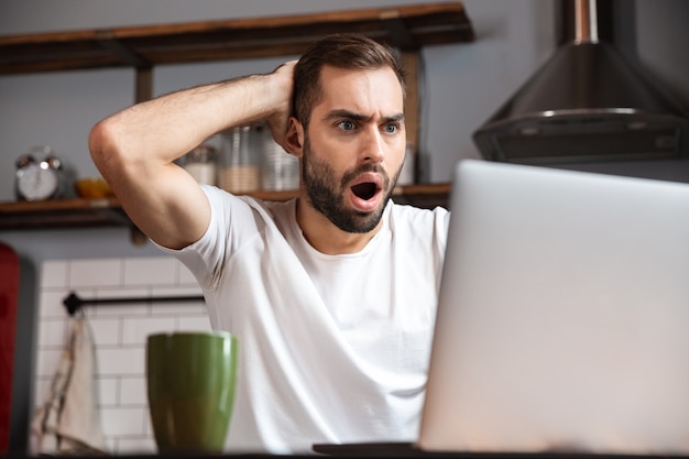 Shocked young man using laptop computer while sitting at the kitchen table