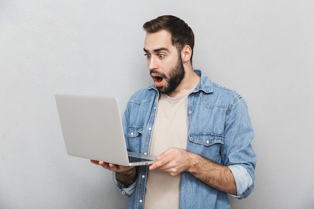 Shocked young man wearing shirt isolated over gray wall, showing laptop computer