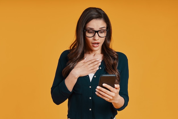 Shocked young woman in casual clothing using smart phone while standing against yellow background