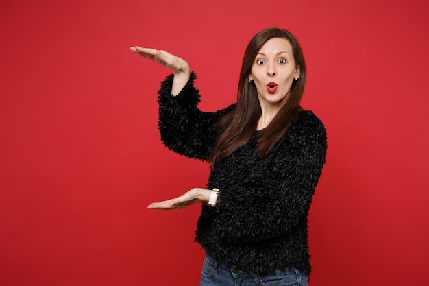 Shocked young woman in fur sweater gesturing demonstrating size with vertical workspace isolated on bright red wall background in studio. People sincere emotions lifestyle concept. Mock up copy space.