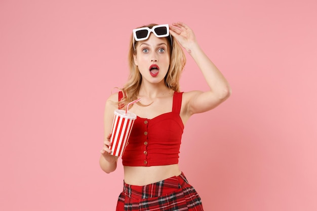 Shocked young woman girl in red sexy clothes, dark sunglasses isolated on pastel pink wall background. People sincere emotions lifestyle concept. Mock up copy space. Hold plastic cup of cola or soda.