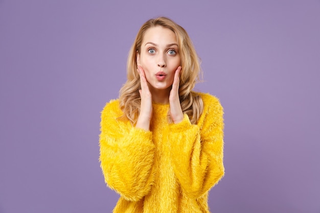 Shocked young woman girl in yellow fur sweater posing isolated on pastel violet wall  portrait. People sincere emotions lifestyle concept.  Put hands on cheeks.