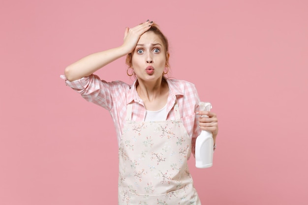 Shocked young woman housewife in casual clothes, apron hold atomizer spray with washing cleanser doing housework isolated on pink background studio portrait. Housekeeping concept. Put hand on head.