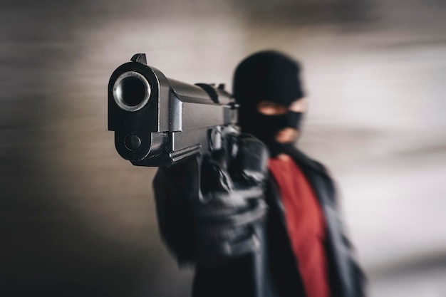 Shooter in black mask attacks concept of banditry theft crime Armed terrorist thief with pistol in hands criminal targeting with gun Mafia gangster point gun in camera Focus on pistol barrel