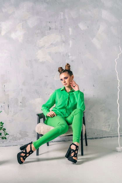 Shooting professional Fashion model in modern studio. Stylish woman in green suit sits on chair on gray background. Vertical frame.