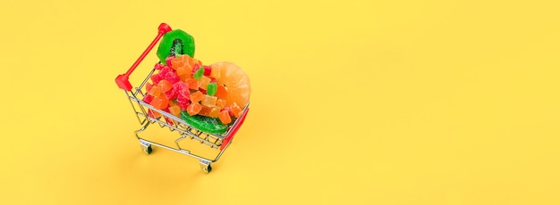 Shopping cart full of colorful colored candied fruit over yellow wall