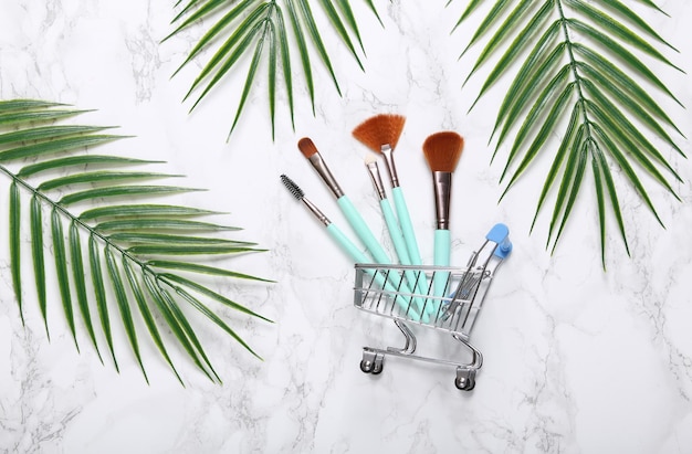 Shopping cart with makeup brushes and palm leaves on marble background