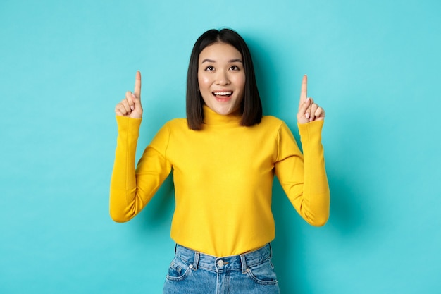 Shopping concept. Beautiful asian woman in yellow sweater pointing fingers up at logo, looking at promotion with happy smile, standing over blue.