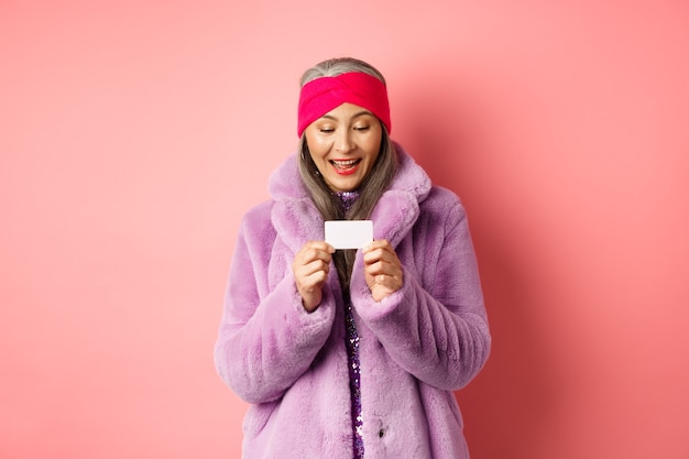 Shopping and fashion concept. Beautiful and stylish asian woman looking at plastic credit card, smiling pleased, standing over pink background.