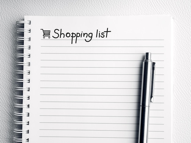 Shopping list handwriting note text on empty notebook page Template white paper sheet with line and shopping cart icon on spiral notepad with a silver pen on white leather background