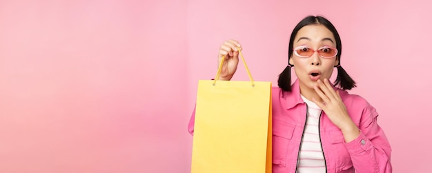 Shopping Stylish asian girl in sunglasses showing bag from shop and smiling recommending sale promo in store standing over pink background