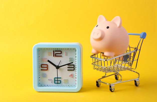 Shopping trolley with piggy bank and Clock on yellow background Shopping time