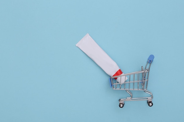 Shopping trolley with tube of cream or toothpaste against blue background. Top view