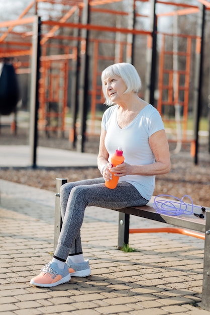 Short break. Cheerful blond woman holding a bottle of juice and relaxing while exercising