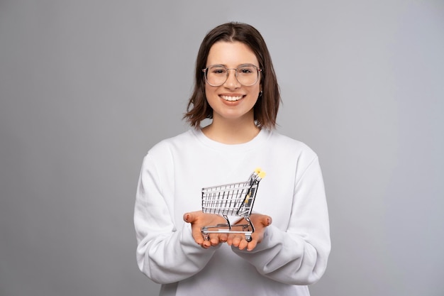 Short haired young woman is holding a small shopping cart while smiling Studio portrait over grey background