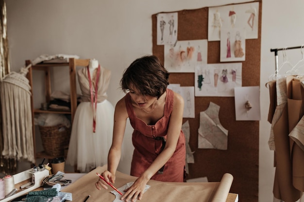 Shorthaired brunette woman in linen red dress poses in cozy office Fashion designer draws cloth pattern on craft paper sheet