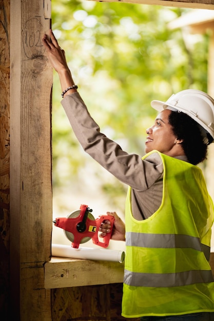 Shot of an African American female architect measuring a window while checking construction site of a new wooden house.