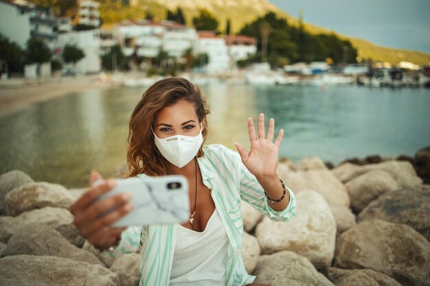 Photo shot of an attractive happy young woman wearing a protective n95 mask and making video call with her smartphone while spending time on the seaside during the covid-19.