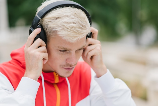 Shot of attractive runner with muscular athletic body dressed in red sportswear resting after physical activities in open air in park Young handsome man listening to music with headphones outdoor