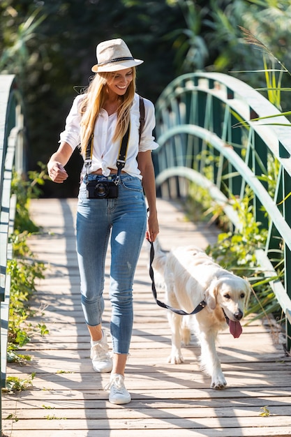 Shot of attractive young amateur photograph woman walking with her dog crossing over a bridge in the park.